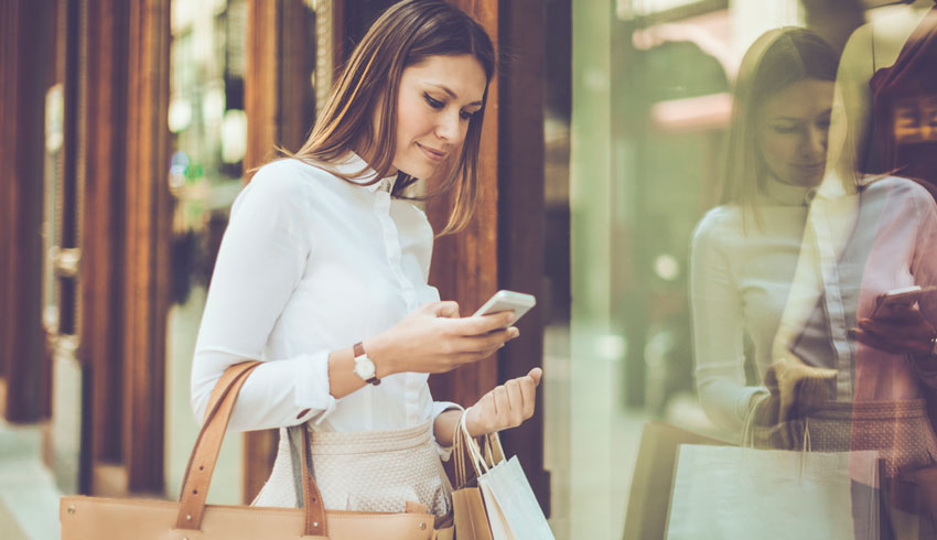 A woman carrying a large purse and shopping bags stops at the front of a store window to check her phone. 